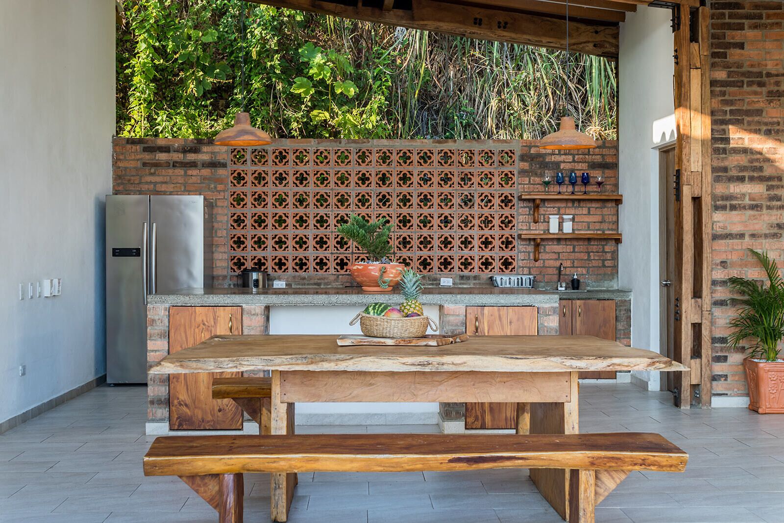 An outdoor kitchen with a wooden table, a refrigerator, and a brick backsplash.