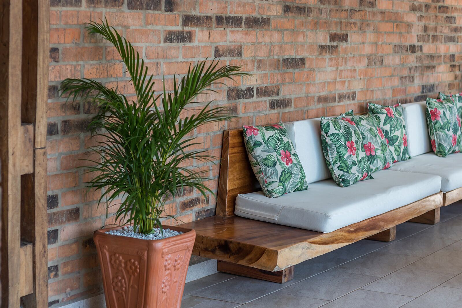 Un banc en bois avec des coussins blancs et des oreillers  à motifs floraux tropicaux, adossé à un mur de briques.