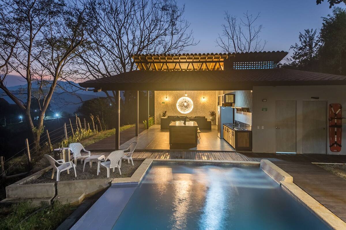 Ein moderner Poolbereich im Freien mit Essbereich und einem durch eine offene Struktur sichtbaren Küchenbereich.