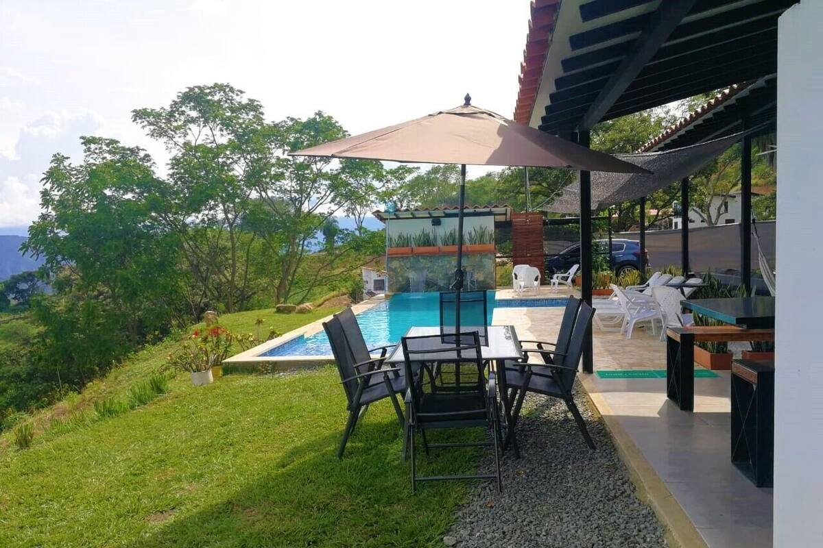 Ein Poolbereich mit Tisch und Stühlen, umgeben von viel Grün und Blick aufs Meer.