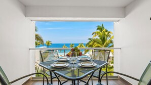 A balcony with a table set for dining, overlooking a pool and palm trees, with the ocean in the background. - Gorgeous 1BR with Pools & Sea Views (Puerto Vallarta)