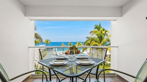 Ein Balkon mit Esstisch, Blick auf Pool und Palmen, im Hintergrund das Meer.