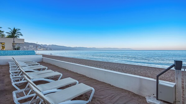 A beachside view with a row of white sun loungers, a tiled pool, and a clear view of the sea and distant mountains.