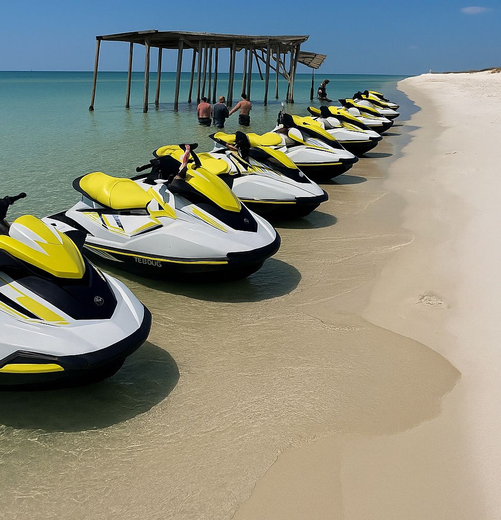 Diversas motos aquáticas alinhadas em uma praia de areia, com um píer de madeira ao fundo.