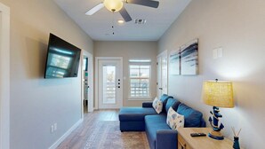 A modern living room with a blue sofa, a wooden coffee table, a floor lamp, and a flat-screen TV mounted on the wall. - Pet Friendly, Shared Heated Pool, Golf Cart Included (Port Aransas)