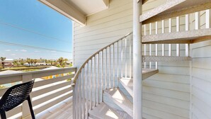 A balcony with a white railing, a black chair, and a view of a residential area with houses and palm trees. - Pet Friendly, Shared Heated Pool, Golf Cart Included (Port Aransas)