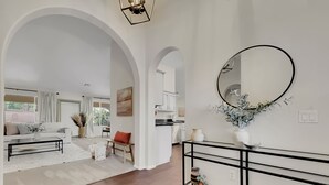 A spacious living area with a white sofa, coffee table, and a dining table with chairs. There is a large mirror on the wall and a vase with flowers on a console table.