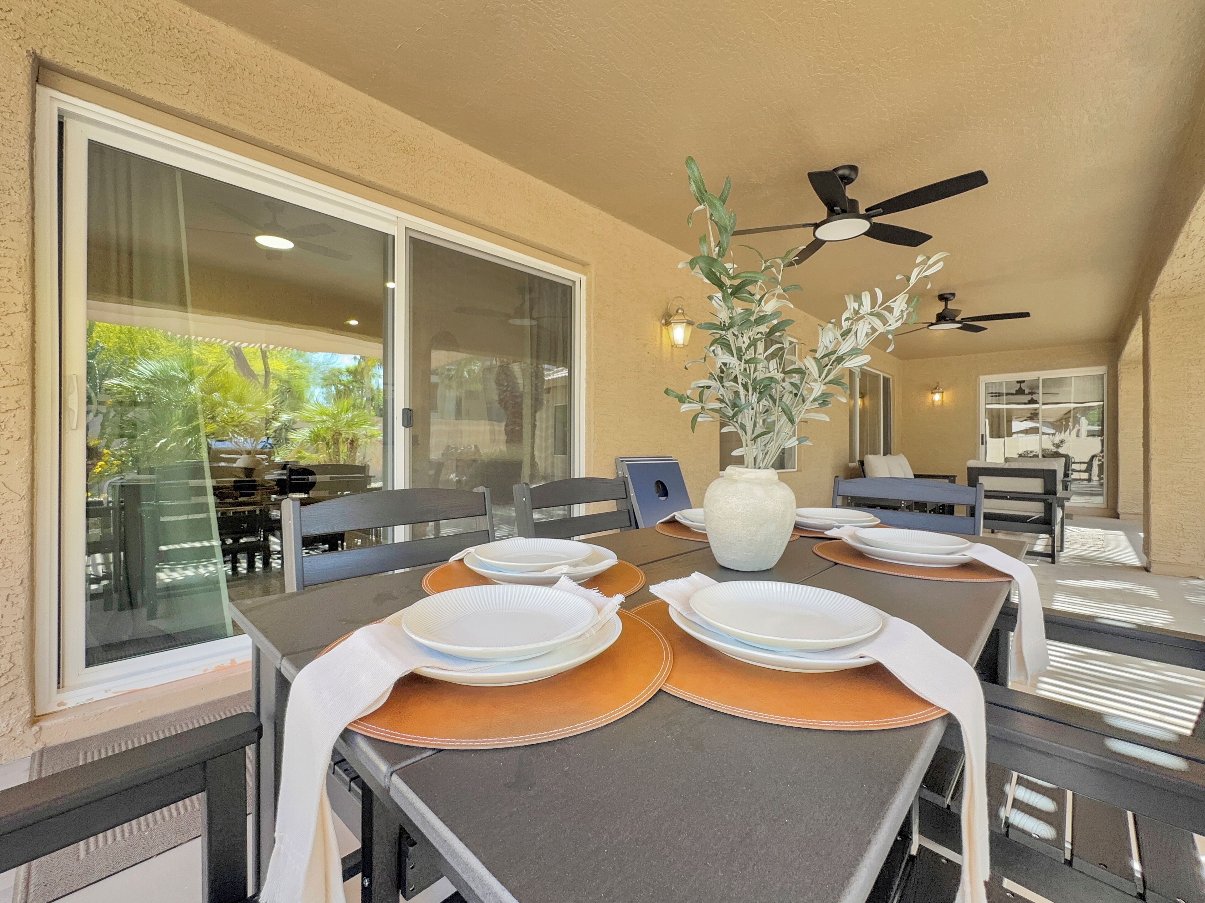 An outdoor dining area with a table set for four, surrounded by chairs, a ceiling fan, and a large window offering a view of the outdoors.