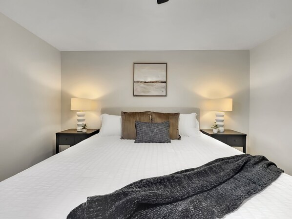 A neatly arranged bedroom with a large bed, two bedside tables with lamps, a framed artwork above the bed, and a dark throw blanket on the bed.