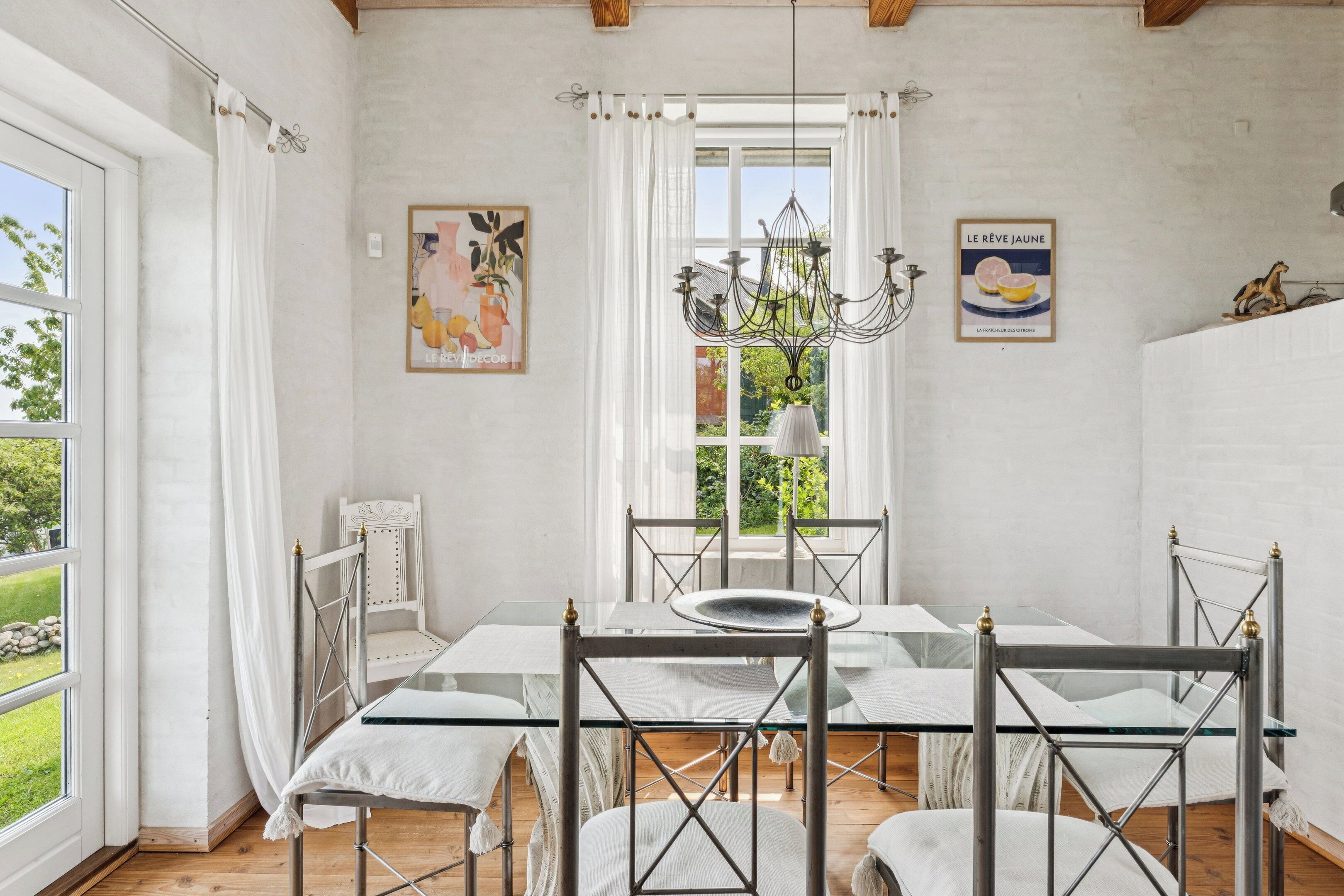 Un espace repas avec une table en verre, des chaises en métal, des rideaux blancs, un lustre et une vue sur la verdure à l’extérieur.