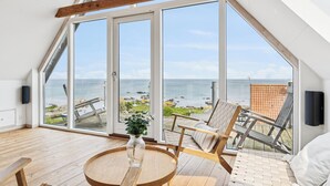 A high-ceilinged room with large windows, a wooden floor, a round table, and a view of the sea. - Luxury holiday home with sea view (Svaneke)