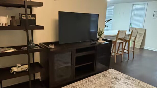 Una sala de estar con un mueble multimedia oscuro, un televisor de pantalla plana, un tapete estampado y una zona de comedor con sillas de madera y una mesa.