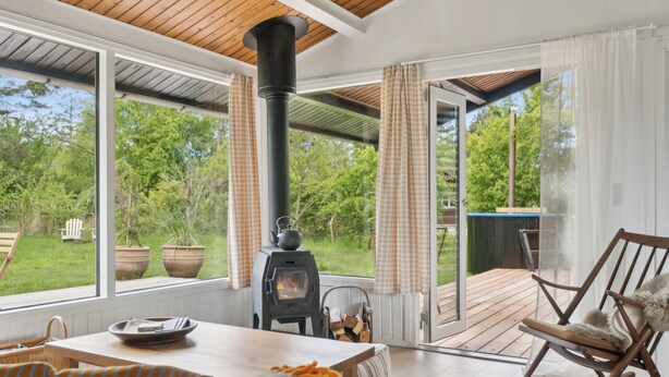 A cozy room with a large window, a fireplace, a rocking chair, and a wooden table. - Cozy holiday home with wood-fired tub and sauna (Rødby)