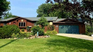 Une maison en bois à deux étages, dotée d’un garage, entourée d’une pelouse bien entretenue et de divers arbustes.