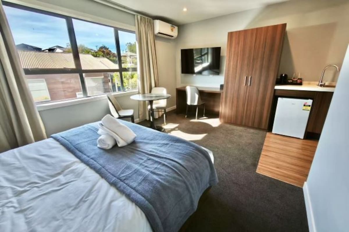 A hotel room with a bed, a desk, a chair, a TV, and a kitchenette.