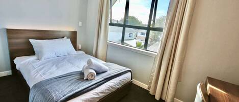 A neatly made bed with a blue and white striped blanket, a wooden headboard, and a view of a building through a window with curtains.