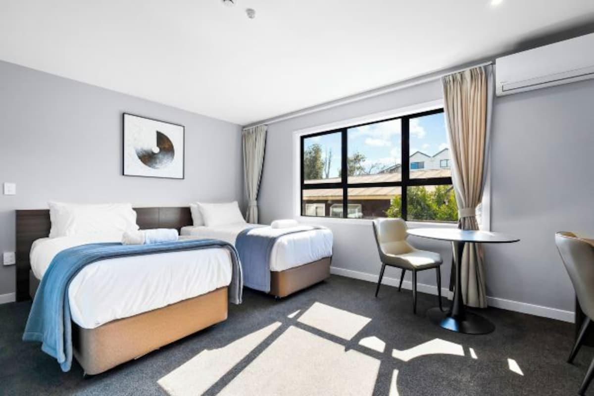 A hotel room with two beds, a desk, a chair, and a large window with curtains.