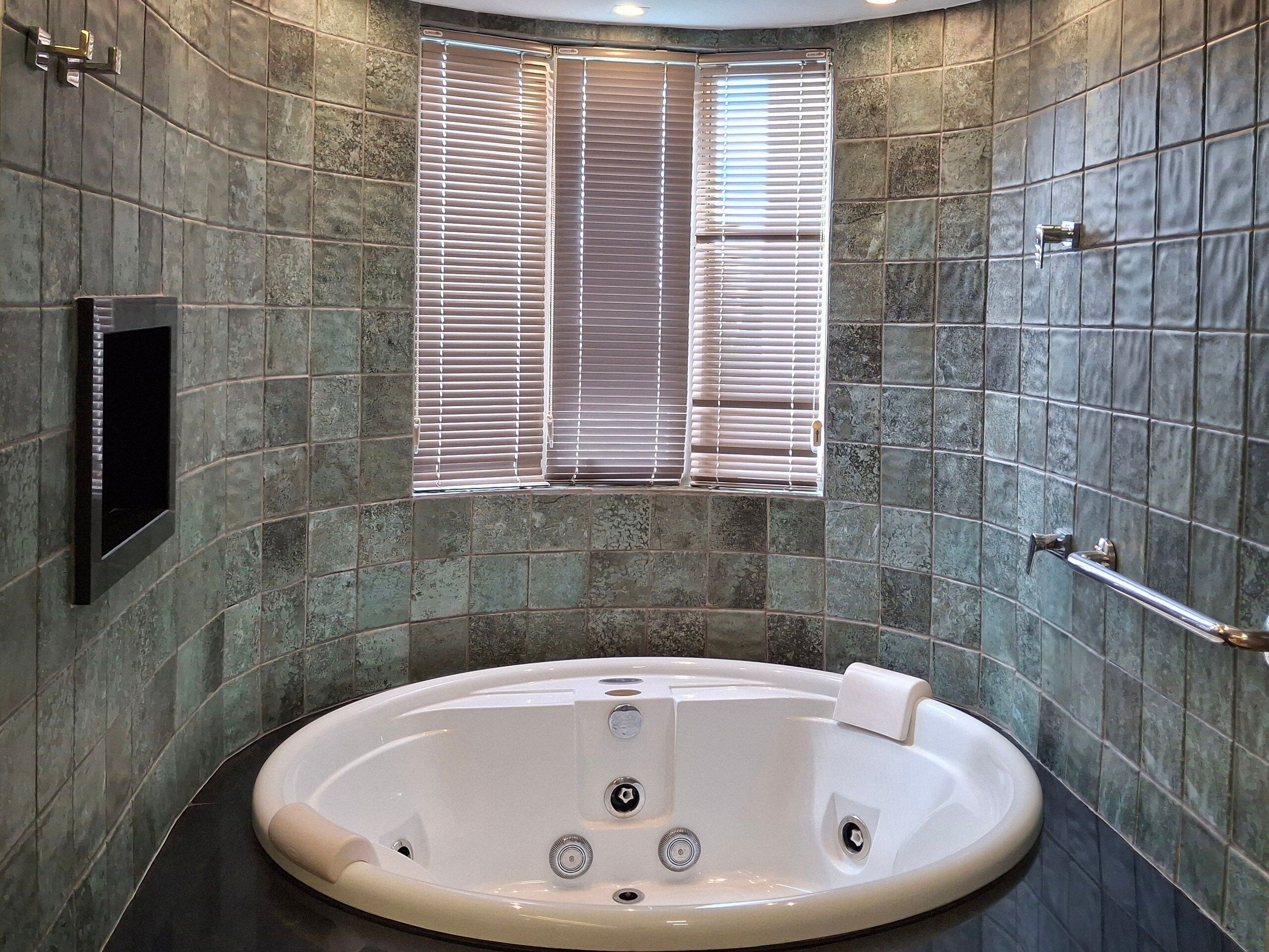 Une salle de bain moderne avec une baignoire sur pieds, une fenêtre avec stores et un téléviseur fixé au mur.