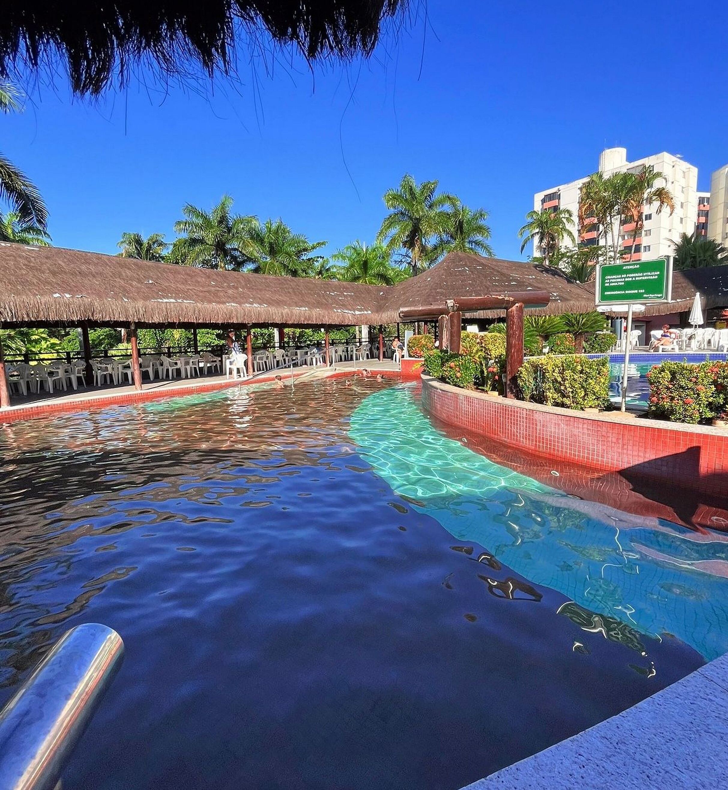 A swimming pool with clear water, surrounded by lounge chairs and umbrellas, with palm trees and a hotel building in the background.