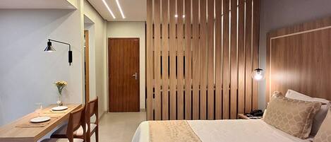 A hotel room with a wooden headboard, a bed with patterned bedding, a desk with a chair, a nightstand, and a wall-mounted lamp.