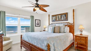 A bedroom with a large bed, a ceiling fan, a view of the ocean, a nightstand with a lamp, and a chair.