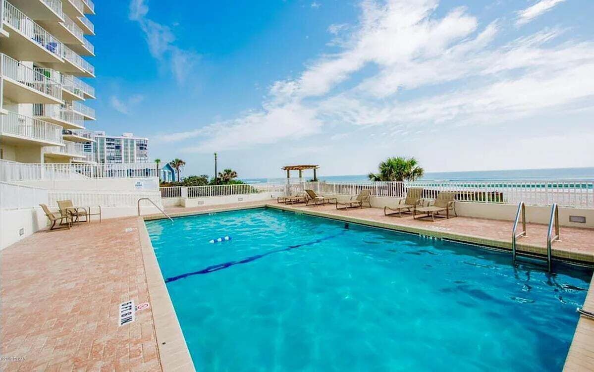 A swimming pool with lounge chairs and a gazebo, surrounded by a white fence, with a view of the ocean and a building with balconies.
