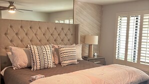 A bedroom with a tufted headboard, a large bed with patterned bedding, a ceiling fan, and a nightstand with a lamp.