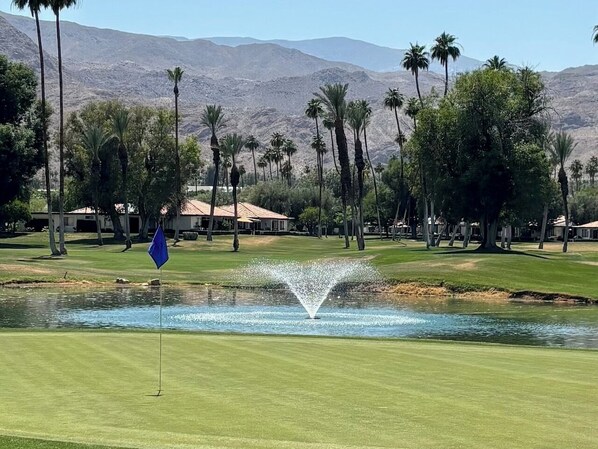 Un campo de golf con una fuente, palmeras y montañas al fondo.