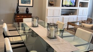 A modern living room with a glass-top dining table, white leather chairs, a wooden cabinet, a television, and decorative items.