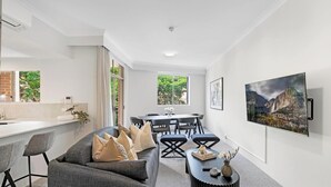 A modern living room with a grey sofa, dining table, and chairs, a kitchen area with a window, and a mounted TV displaying a mountain scene. - KozyGuru | Camperdown | Cozy 2-Bed Apt + Parking (Camperdown)