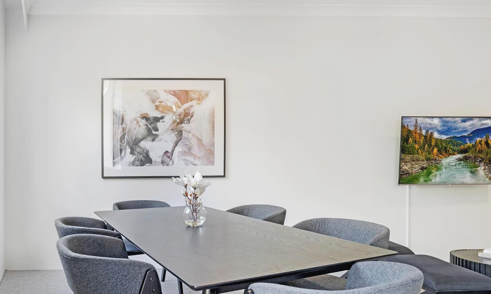 A conference room with a long table, six grey chairs, a vase with white flowers, and two framed artworks on the walls.