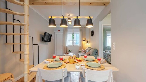 Een eetgedeelte met een houten tafel gedekt voor vier personen, omringd door witte stoelen. Er zijn hanglampen, een televisie aan de muur en een raam met gordijnen.