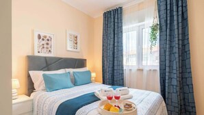 Een net opgemaakt bed met een blauw-witte sprei, twee kussens en een dienblad met ontbijtartikelen zoals croissants en fruit.