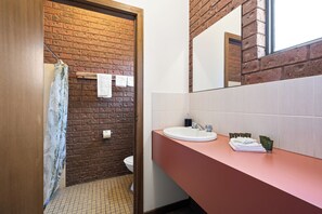 A bathroom with a brick wall, a white toilet, a sink with a mirror, and a shower curtain. - Hotham Motel (Harrietville)