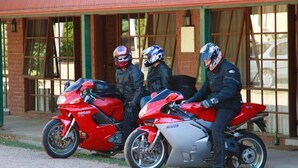 Tres motociclistas con sus motocicletas estacionadas frente a un edificio de grandes ventanales y fachada de ladrillo.