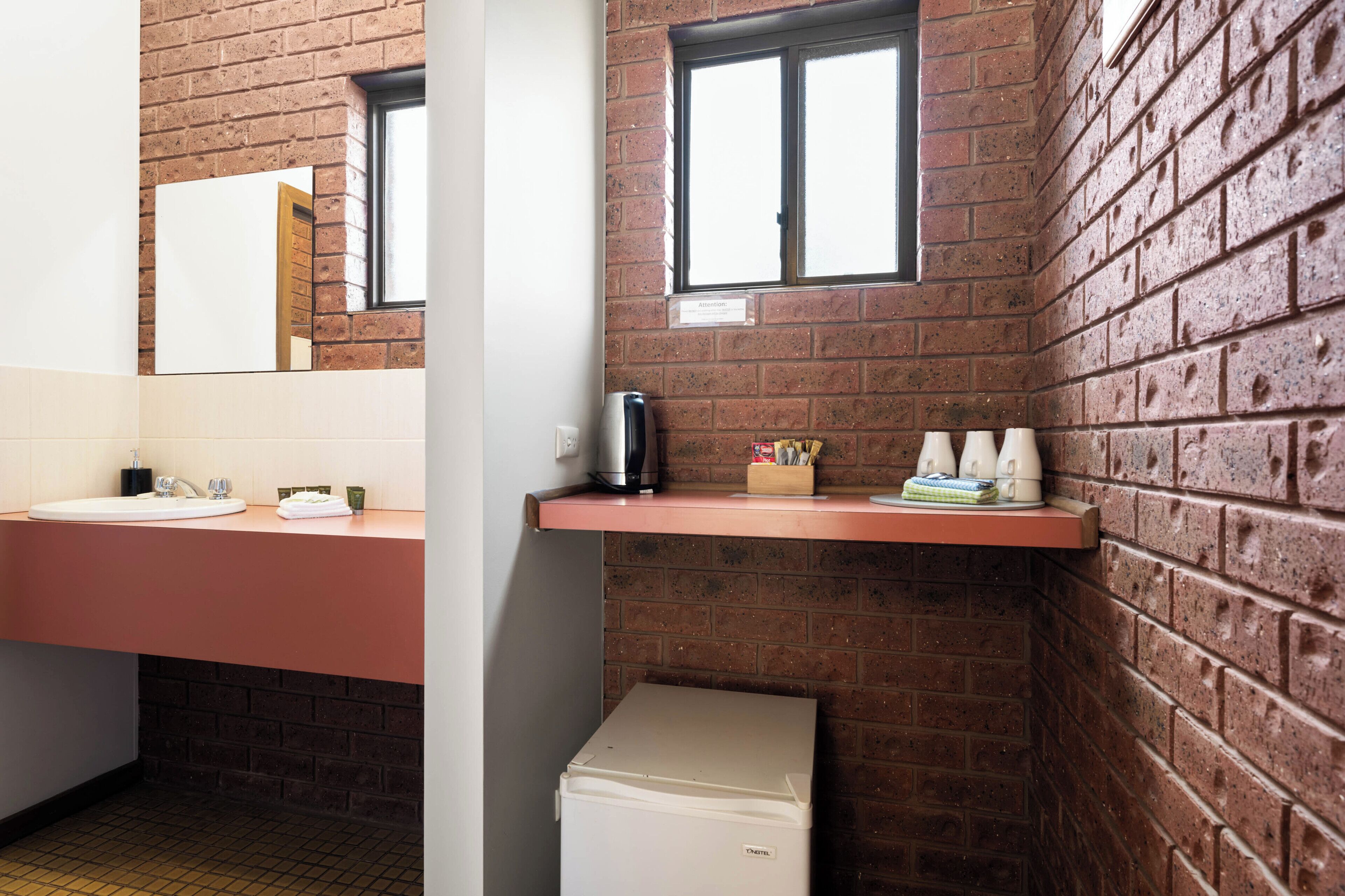 A bathroom with a brick wall, a sink, a mirror, a window, and a small refrigerator.