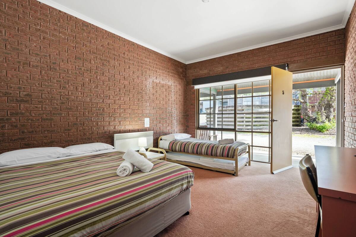 Habitación de hotel con pared de ladrillo, un ventanal grande, una cama con cubrecama a rayas, una mesita de noche y un escritorio.