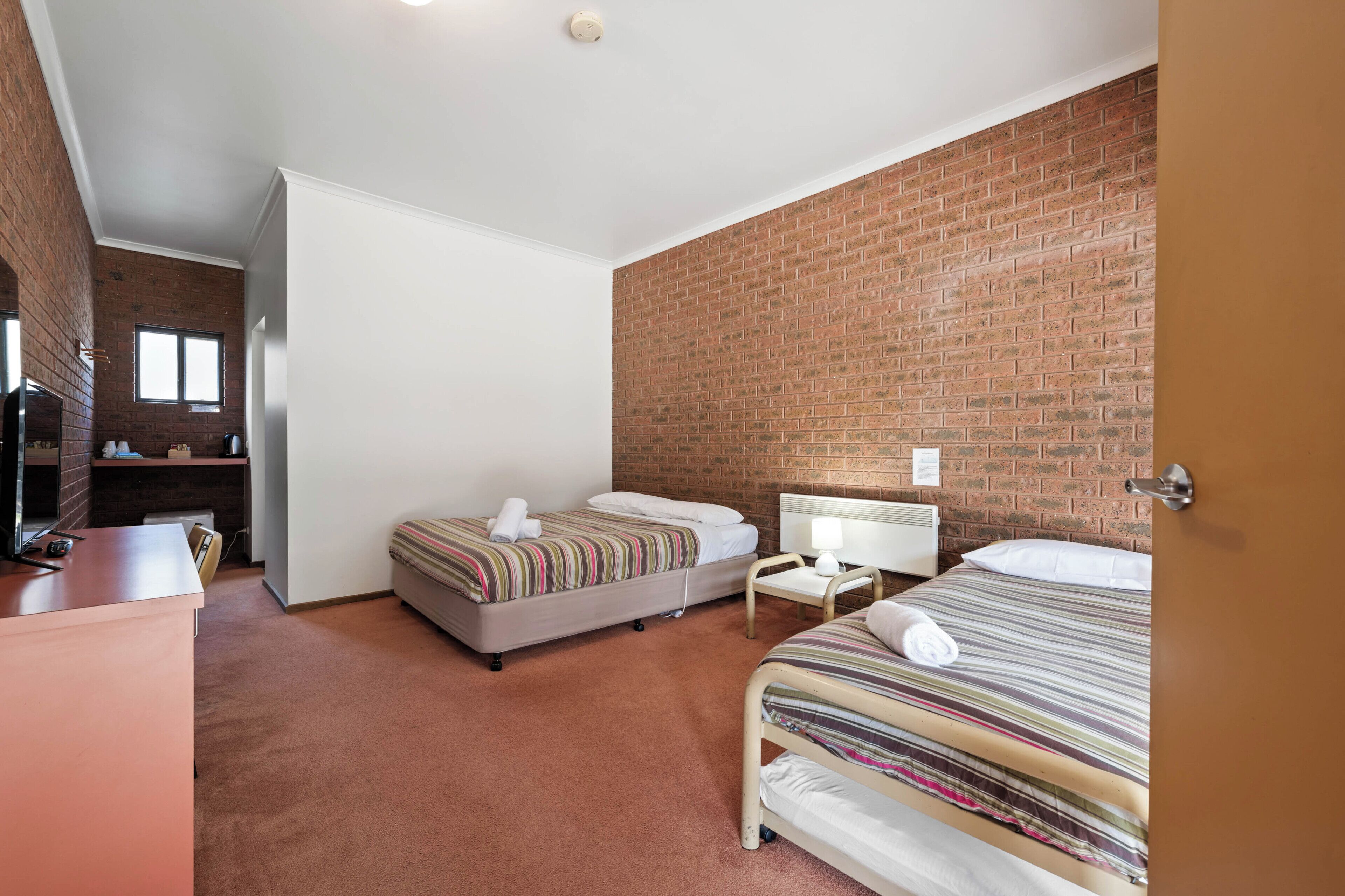 A hotel room with two beds, a desk, a chair, a brick wall, and a white ceiling.