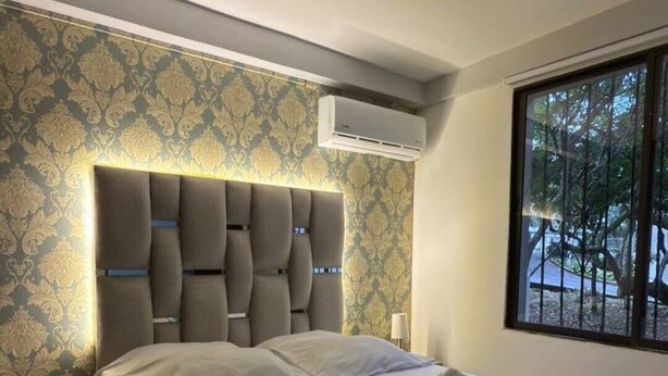 Une chambre moderne avec un grand lit, une tête de lit dotée d’un éclairage intégré, un appareil de climatisation et une fenêtre donnant sur des arbres.