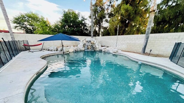 A swimming pool with a stone waterfall feature, a blue umbrella, and a chair. - Spacious 3-bedroom house with pool in a peaceful area in Bakersfield. (Bakersfield)