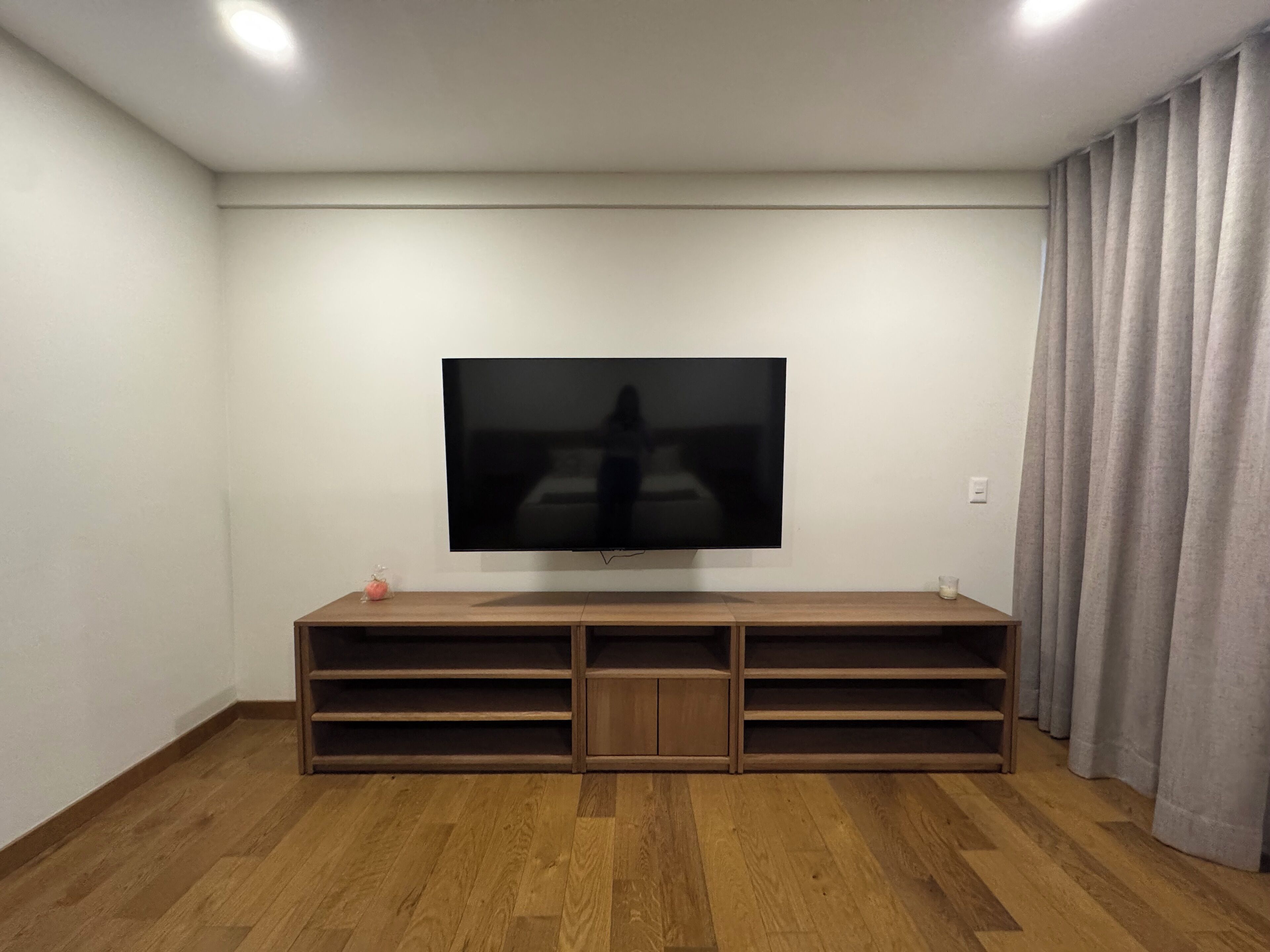 A room with a large flat-screen TV mounted on a wooden entertainment unit, a wooden floor, and a curtain on the right side.