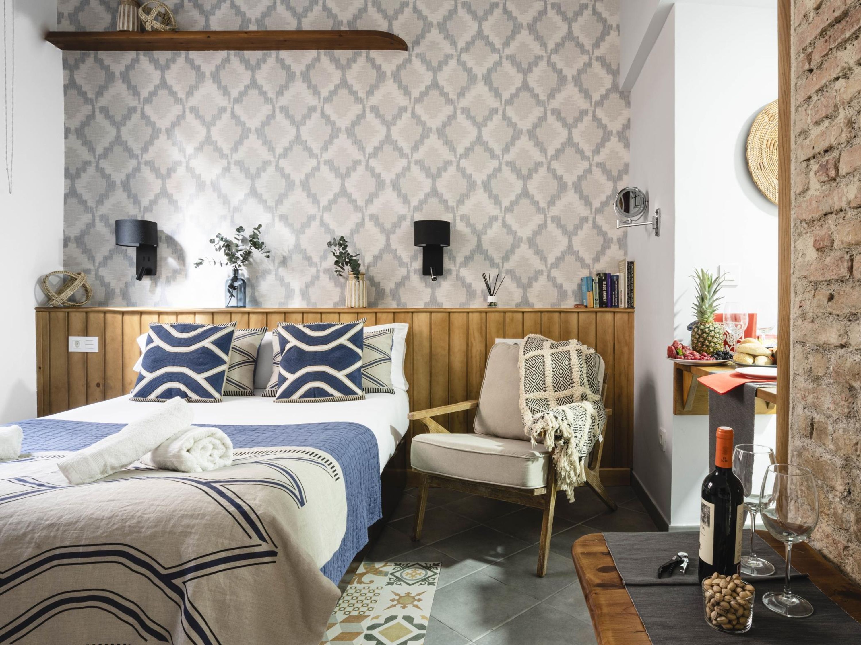 A bedroom with a wooden headboard, patterned wallpaper, a bed with blue and white bedding, a chair with a patterned throw, a small table with a bottle and glasses, and a brick wall.