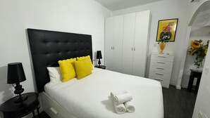 Habitación de hotel con cabecera negra, ropa de cama blanca y almohada de color amarillo. Se ve un aparador blanco con un jarrón de girasoles y un cuadro en la pared.