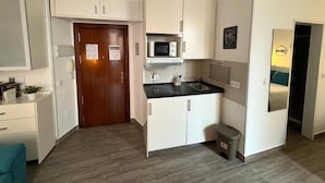 A compact kitchen with white cabinets, a microwave, and a sink. There's a small trash bin and a door leading to another room.