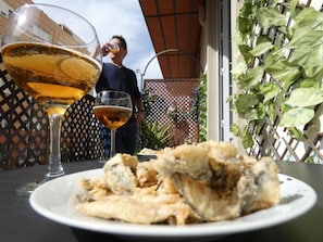 A dining table with a plate of fried fish and two glasses of beer, a man drinking from one of the glasses, and a lattice fence with greenery behind. - Sol de M Laga (Málaga)