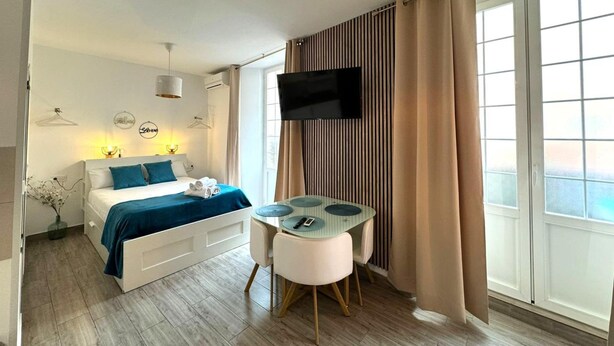 A modern hotel room with a bed, a TV, a small table, and a window with curtains.