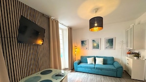 A modern living room with a blue sofa, a glass table, a flat-screen TV mounted on a wooden panel, and a pendant light.