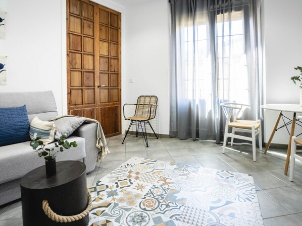 A living room with a grey sofa, a black coffee table, a patterned rug, a wooden door, and a window with grey curtains. - 7 Condes 4 (Málaga)