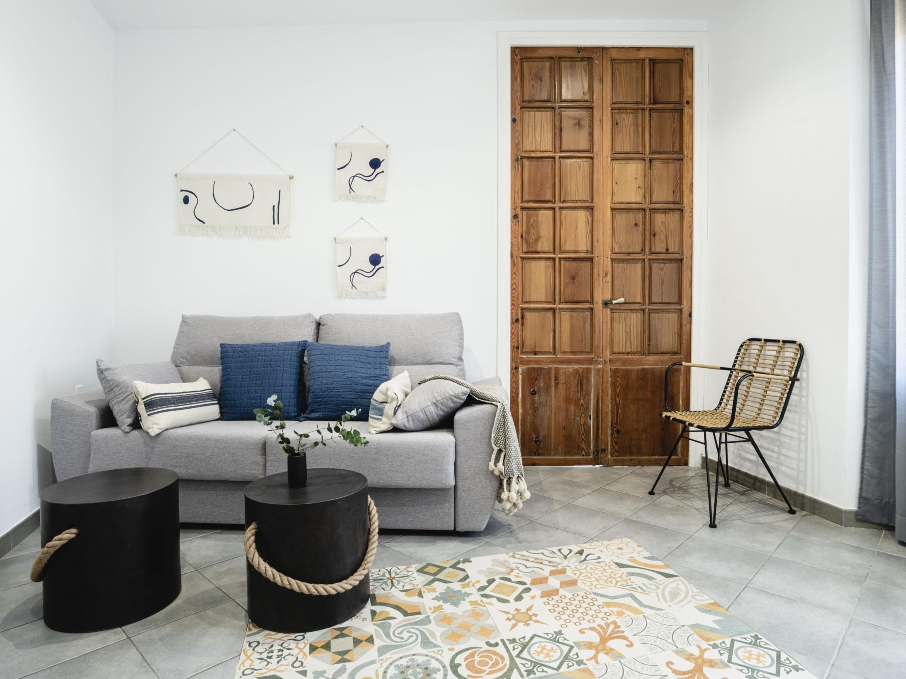 Ein Wohnzimmer mit grauem Sofa, zwei schwarzen Beistelltischen, einem gemusterten Teppich und einer Holztür.