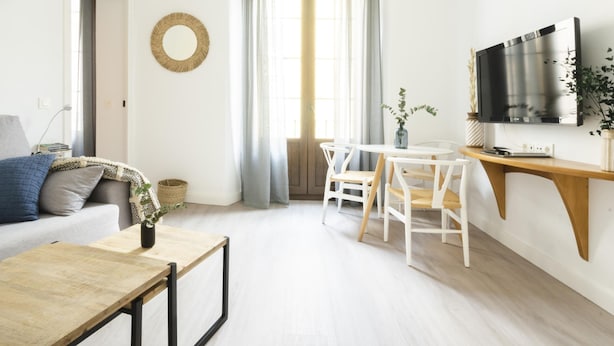 Ein modernes Wohnzimmer mit einer Couch, einem Couchtisch, einem Esstisch mit Stühlen, einem an einem Holzgestell befestigten Fernseher und einem großen Fenster mit Vorhängen.